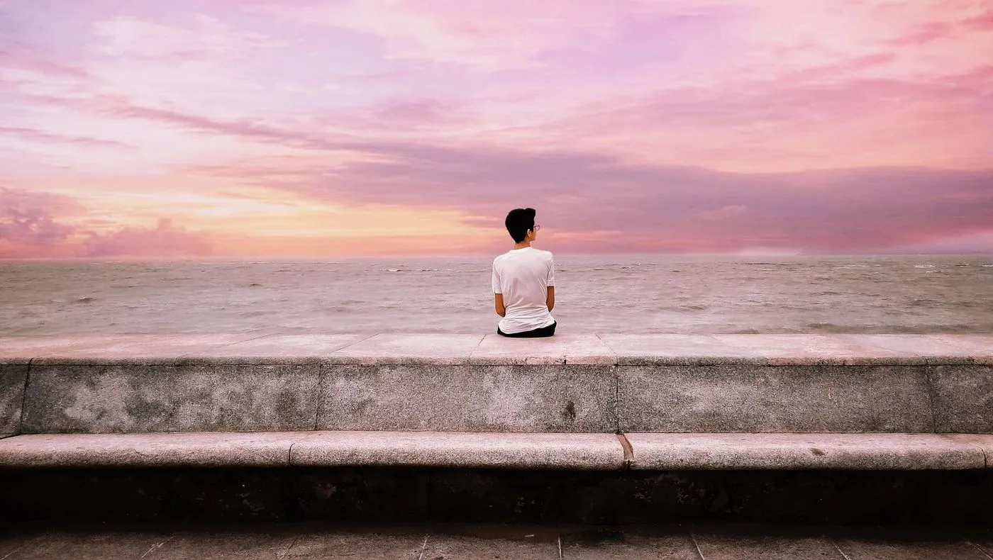 A person sitting alone facing the water at dusk