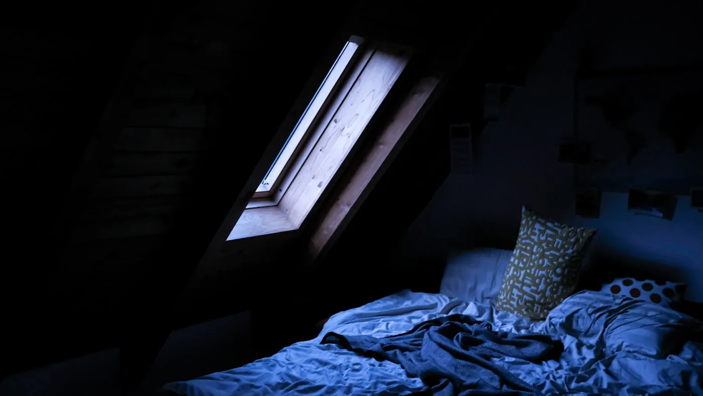 An unmade bed under a skylight at night